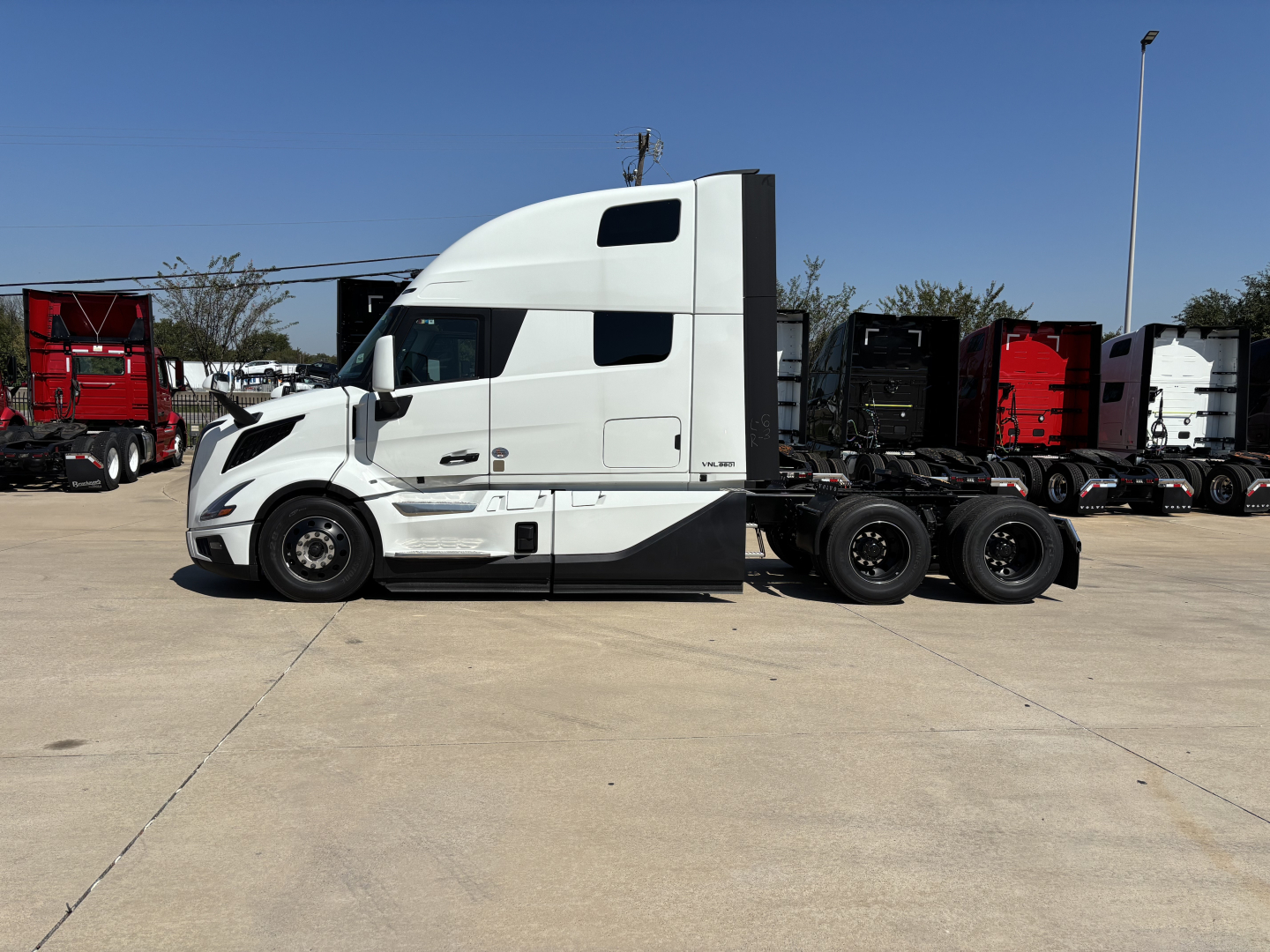 Side view of a white Volvo VNL64T860 tractor-trailer truck with sleeper cab, parked on a lot with other trucks in background.