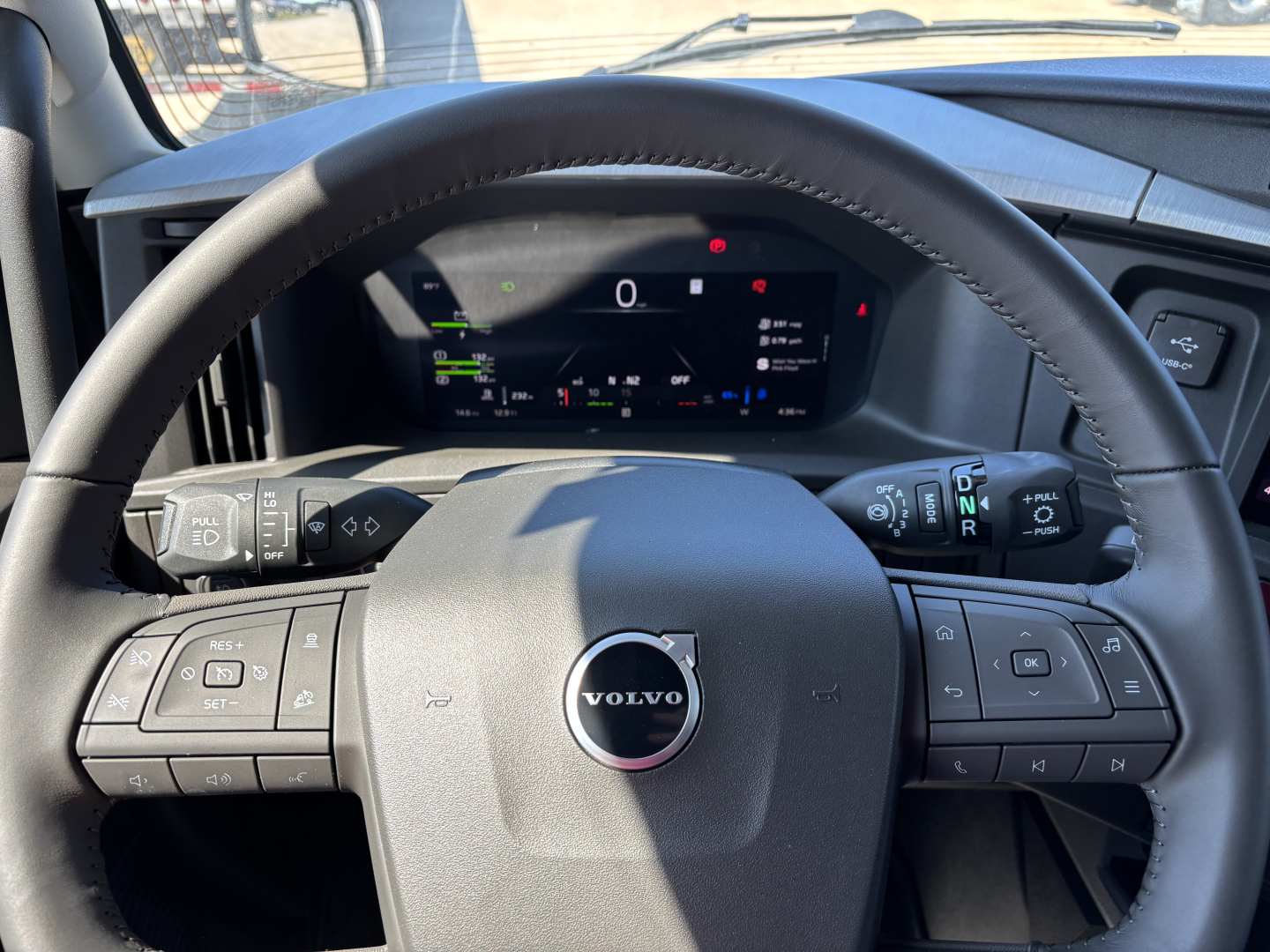 Volvo truck steering wheel and digital instrument cluster in view showing steering wheel controls and Volvo logo.