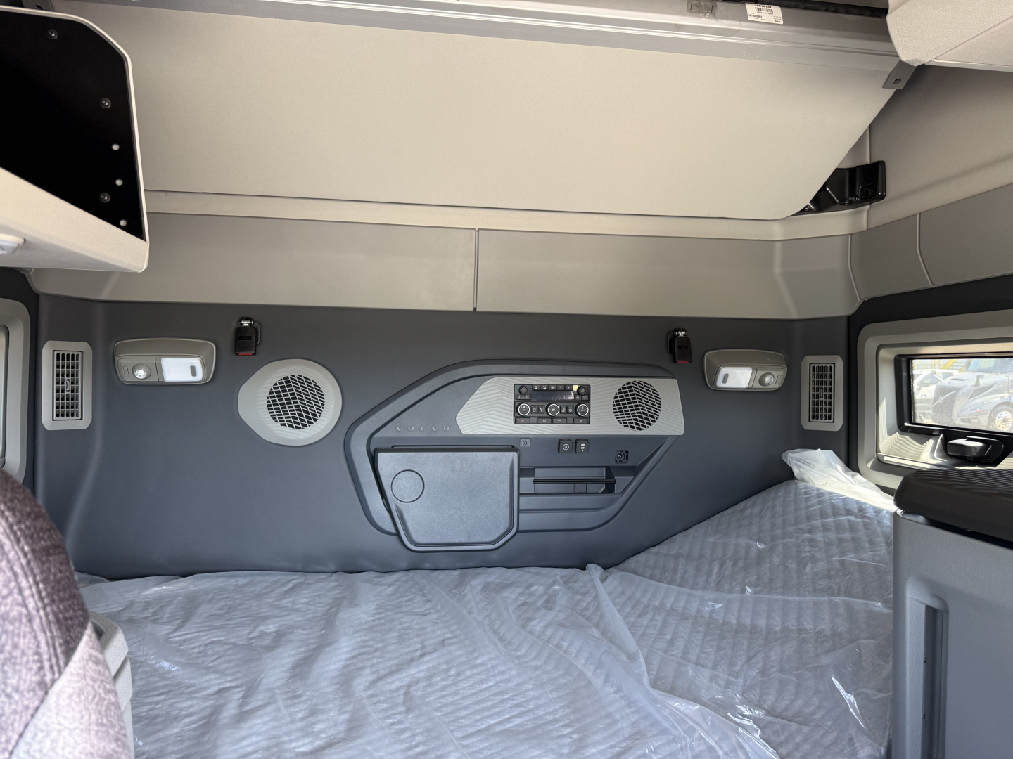 Volvo VNL sleeper cab interior showing control panel, storage cubby, and window, with gray panels and bed in foreground.