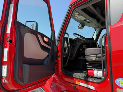 Cab entry of a red Volvo VNL sleeper truck showing open door and interior steering wheel, dashboard, and a fire extinguisher mounted under the dash.