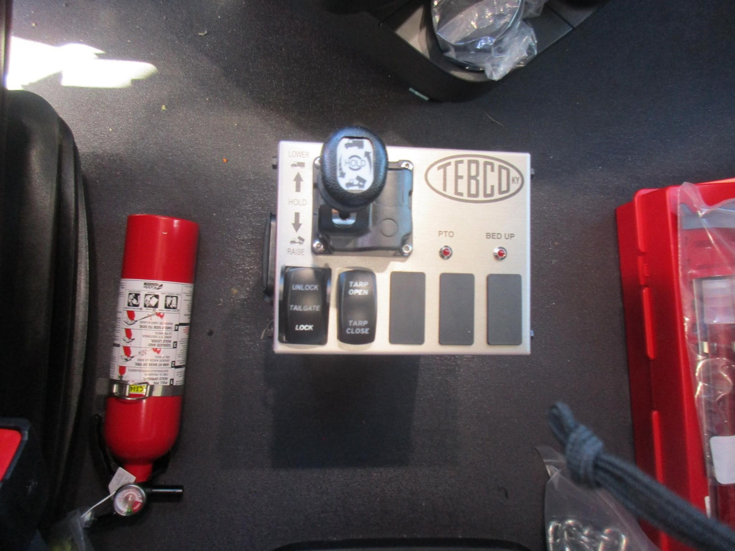 Control panel with TEBCO label and a small gauge, two black lock buttons, and a red fire extinguisher on the left; dashboard area inside a Western Star 47X dump truck cab.
