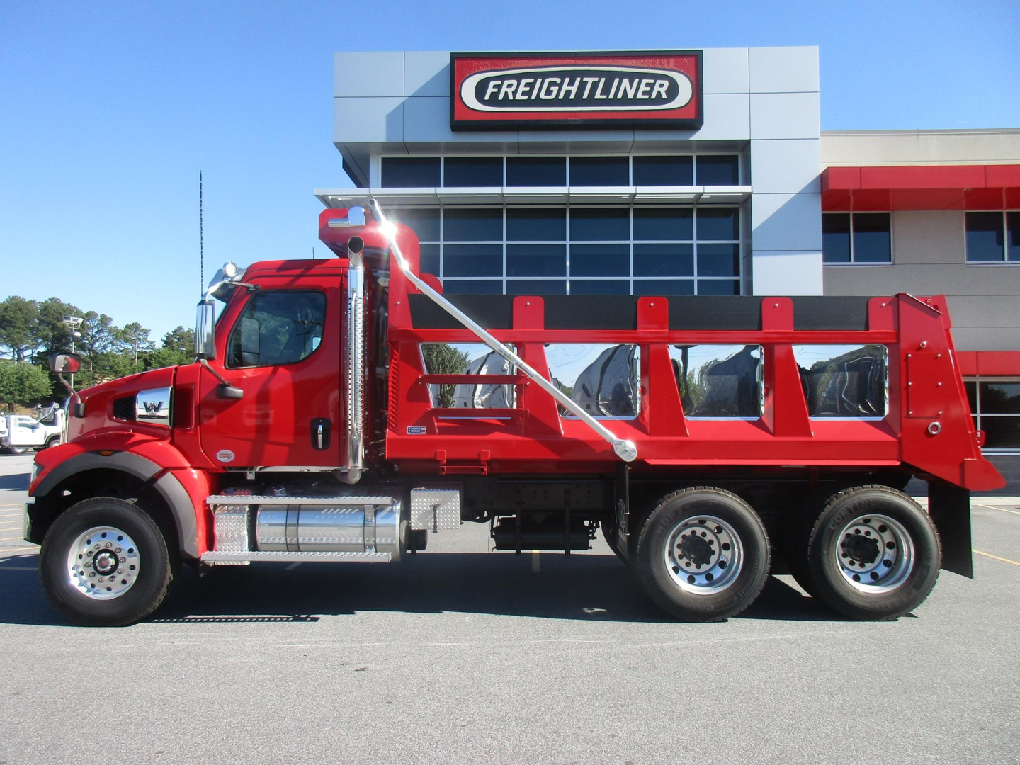 Western Star 47X dump truck in red parked in front of Freightliner dealership building. Side view shows long dump bed, chrome exhaust, dual rear axles, and chrome fuel tanks.