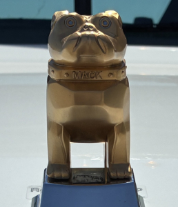 Gold-colored bulldog hood ornament on Mack truck showing close-up of the mascot and base with 'MACK' inscription.