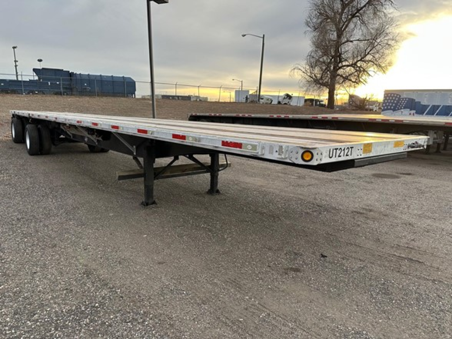 Trailer deck of a flatbed semi-trailer viewed from side, UT2 12T plate visible, metal frame, support legs extended, light bar along edge, parking lot background.