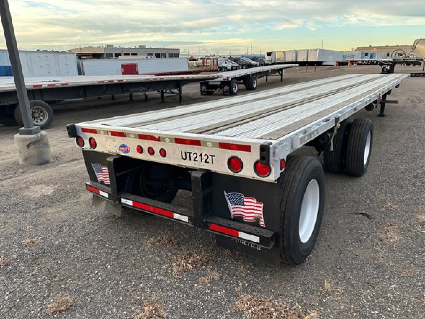 Flatbed trailer showing UT212T license plate area and dual rear axles with white wheels, aluminum deck, rear lights, and bumper with stickers.