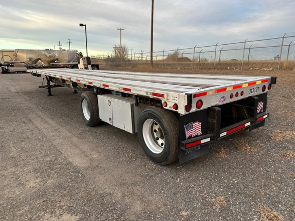 Flatbed trailer with dual rear axles and metal deck, showing Ujack 2 placard and tail lights.