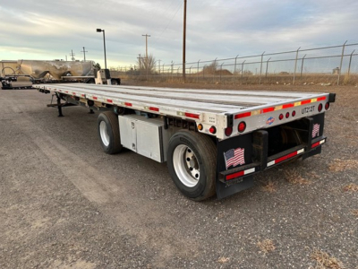 Flatbed trailer with dual rear axles and metal deck, showing Ujack 2 placard and tail lights.