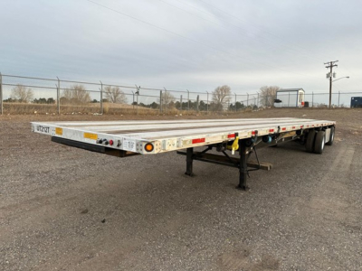 Flatbed trailer in outdoor lot showing long aluminum deck and tandem axles with support legs and rear taillights.