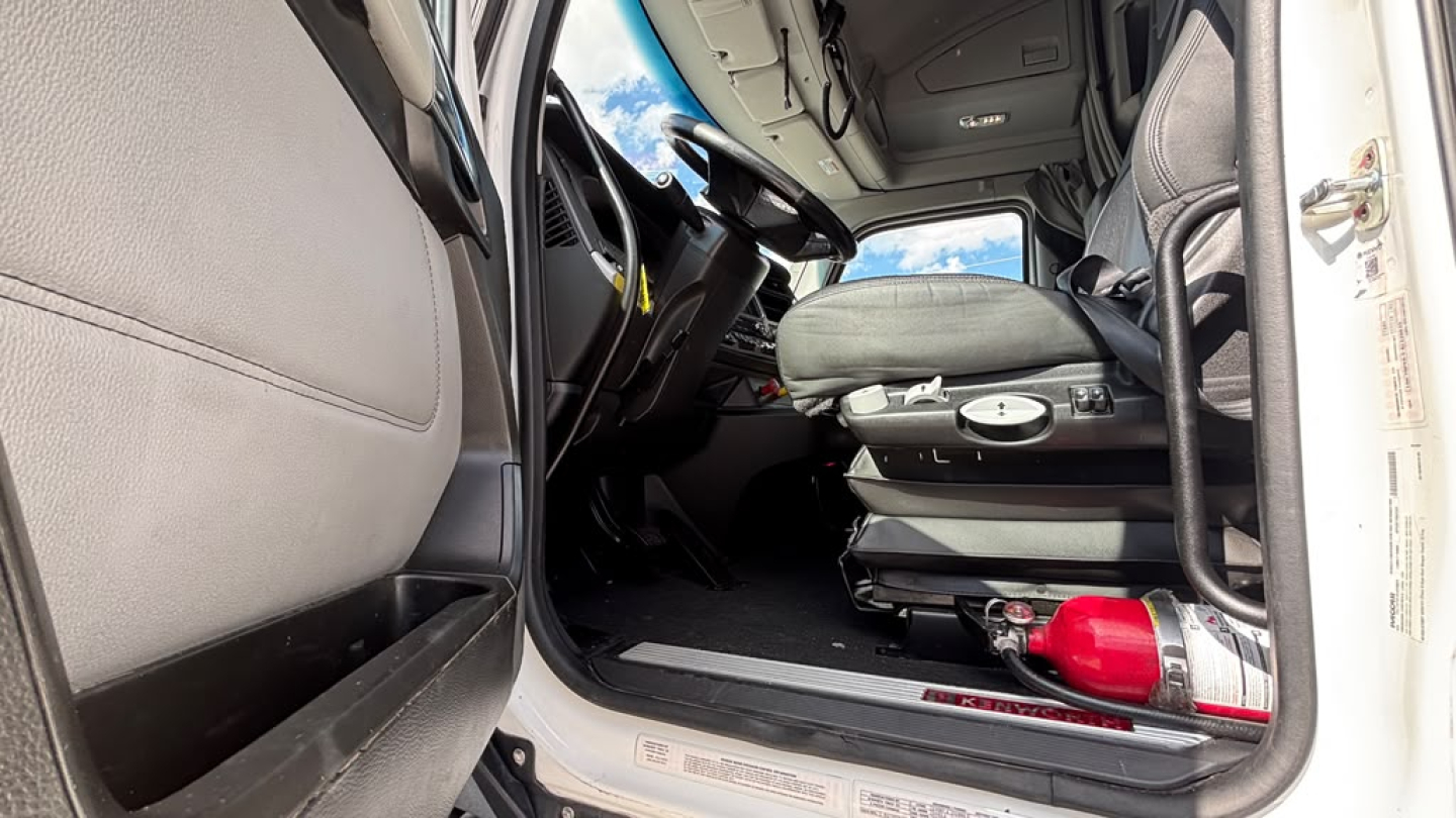 Kenworth T680 cab interior view showing driver seat and dashboard with steering wheel and red fire extinguisher in visibility.
