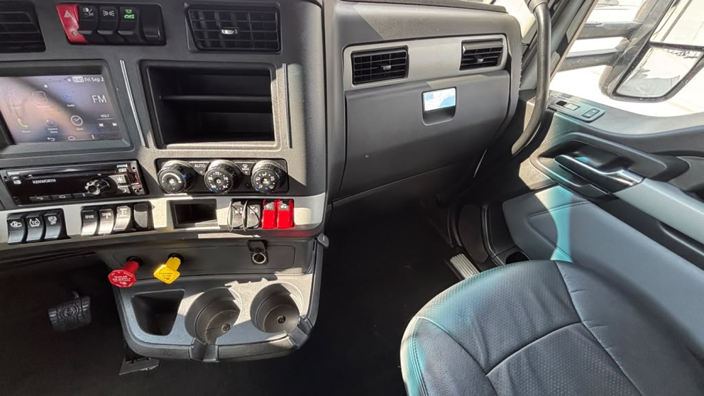 Cab interior of a Kenworth T680 showing dashboard controls, vents, and two cup holders with ignition switches and gauges visible.