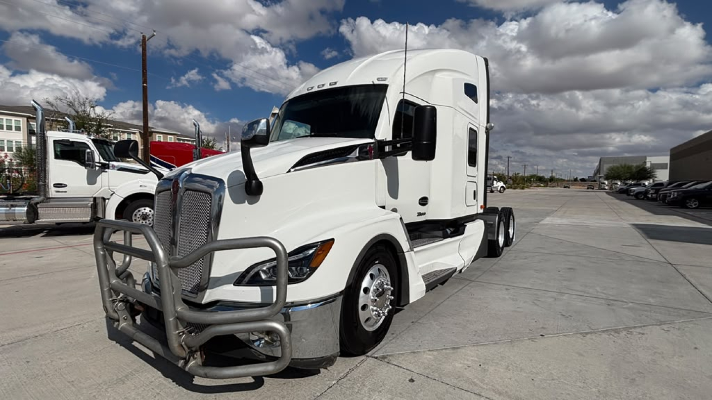 Kenworth T680 conventional sleeper truck in a parking lot, white cab, chrome bumper guard, front grille, and dual rear axles.