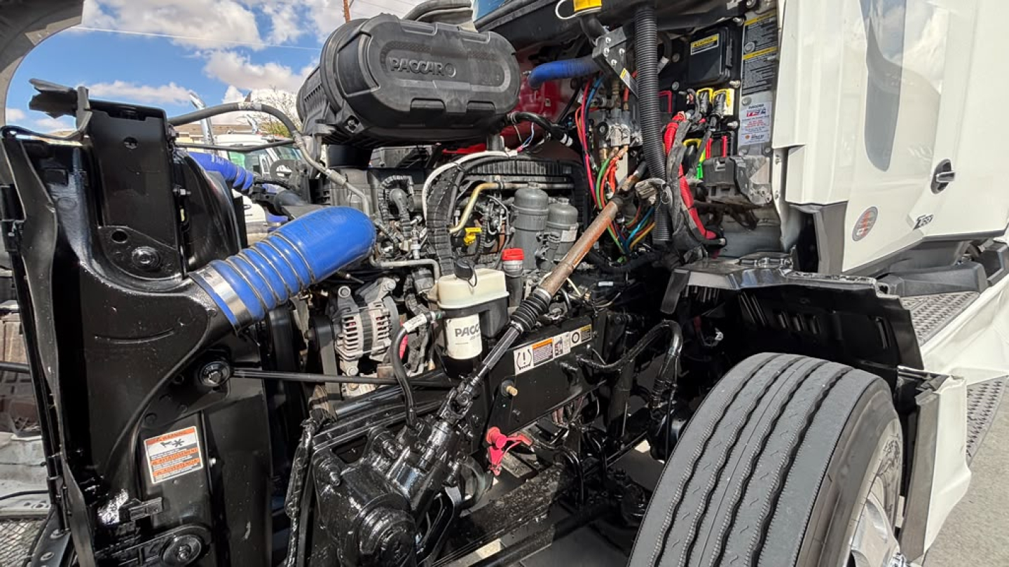 Kenworth T680 engine bay view showing various hoses, wiring, and the diesel engine with blue intake hose and exposed components.
