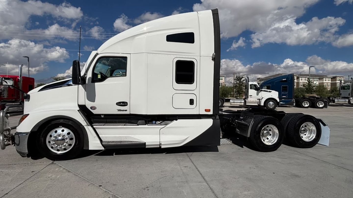 Truck side profile showing a white Kenworth T680 sleeper cab, with black accents and dual rear axles, daytime sky visible.