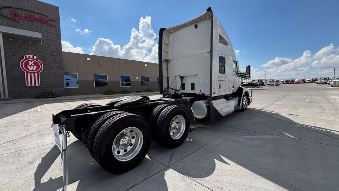 Kenworth T680 tractor in a lot, side view showing cab and rear dual axles, white sleeper cab, dealership building in background; no visible text on components aside from exterior.