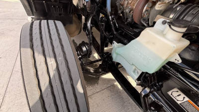 Kenworth T680 front wheel area showing coolant reservoir and engine components with hoses and frame.