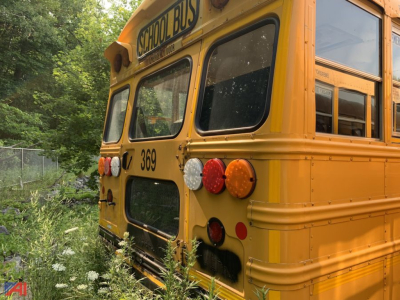School bus rear-left view with yellow body, black bumper, multiple taillights, 'SCHOOL BUS' sign, 369 route number, 2014 Blue Bird Vision