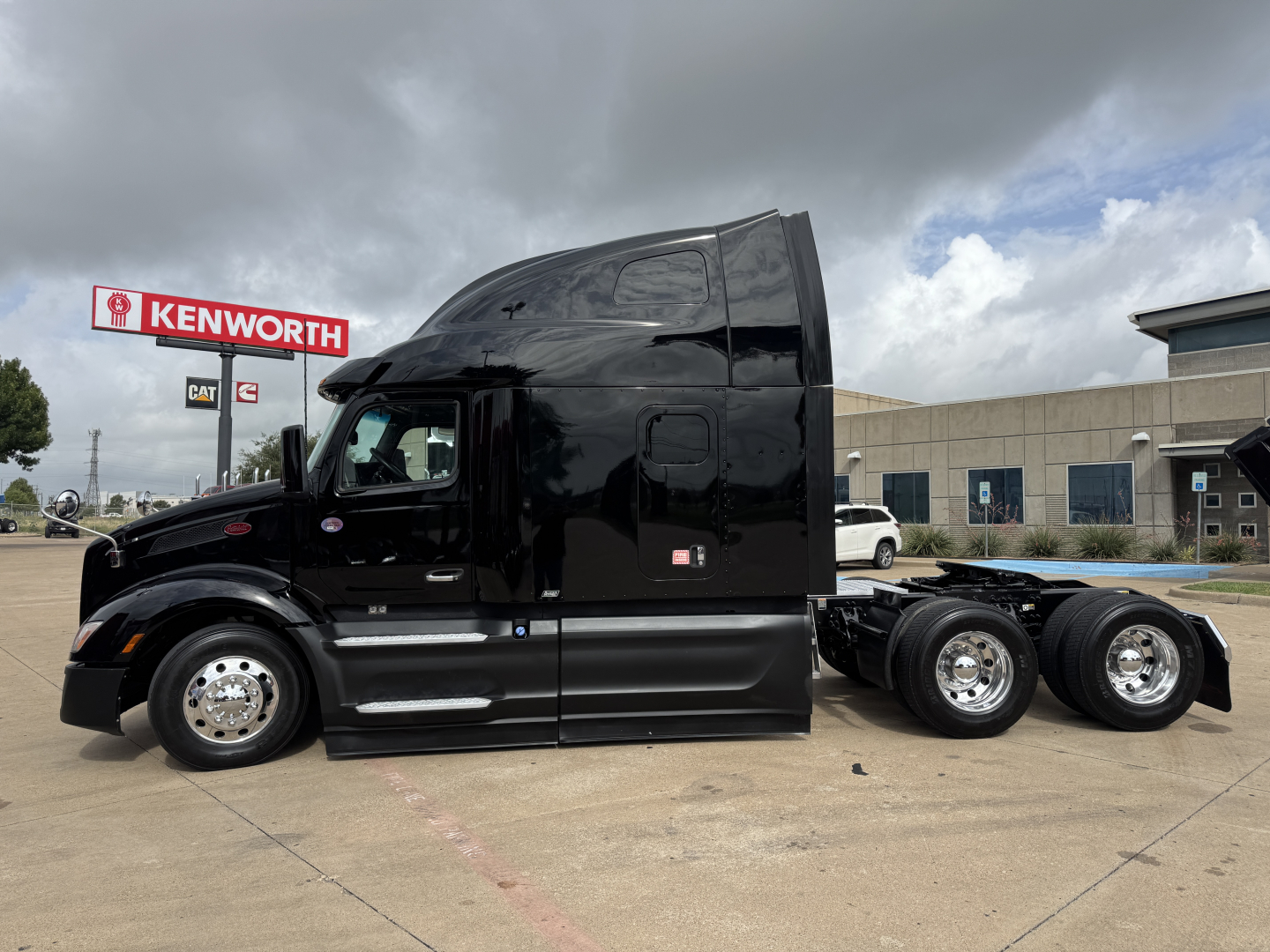 2023 Peterbilt 579 black sleeper tractor side view showing fuel tanks and triple rear axles. Visible text includes Kenworth sign in background and partial license/stock text on listing.