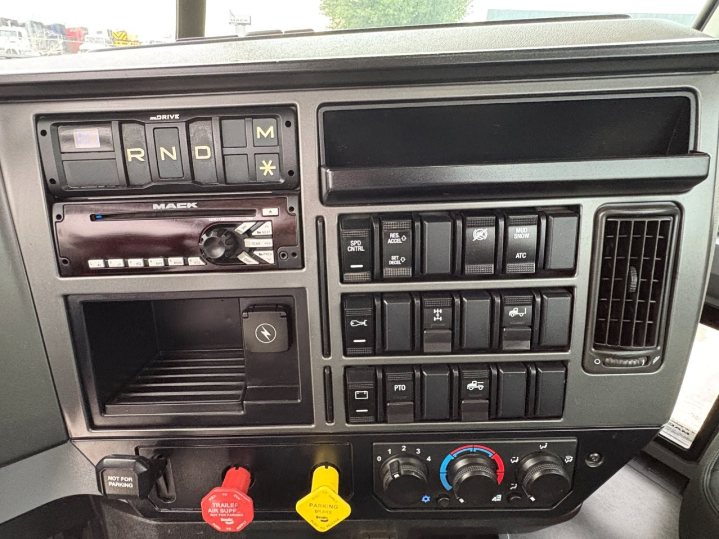 Dashboard of a 2020 Mack PI64T cab showing gear selector with R N D M, stereo, various switches, ignition area, control knobs for climate and auxiliary buttons, and a storage slot.