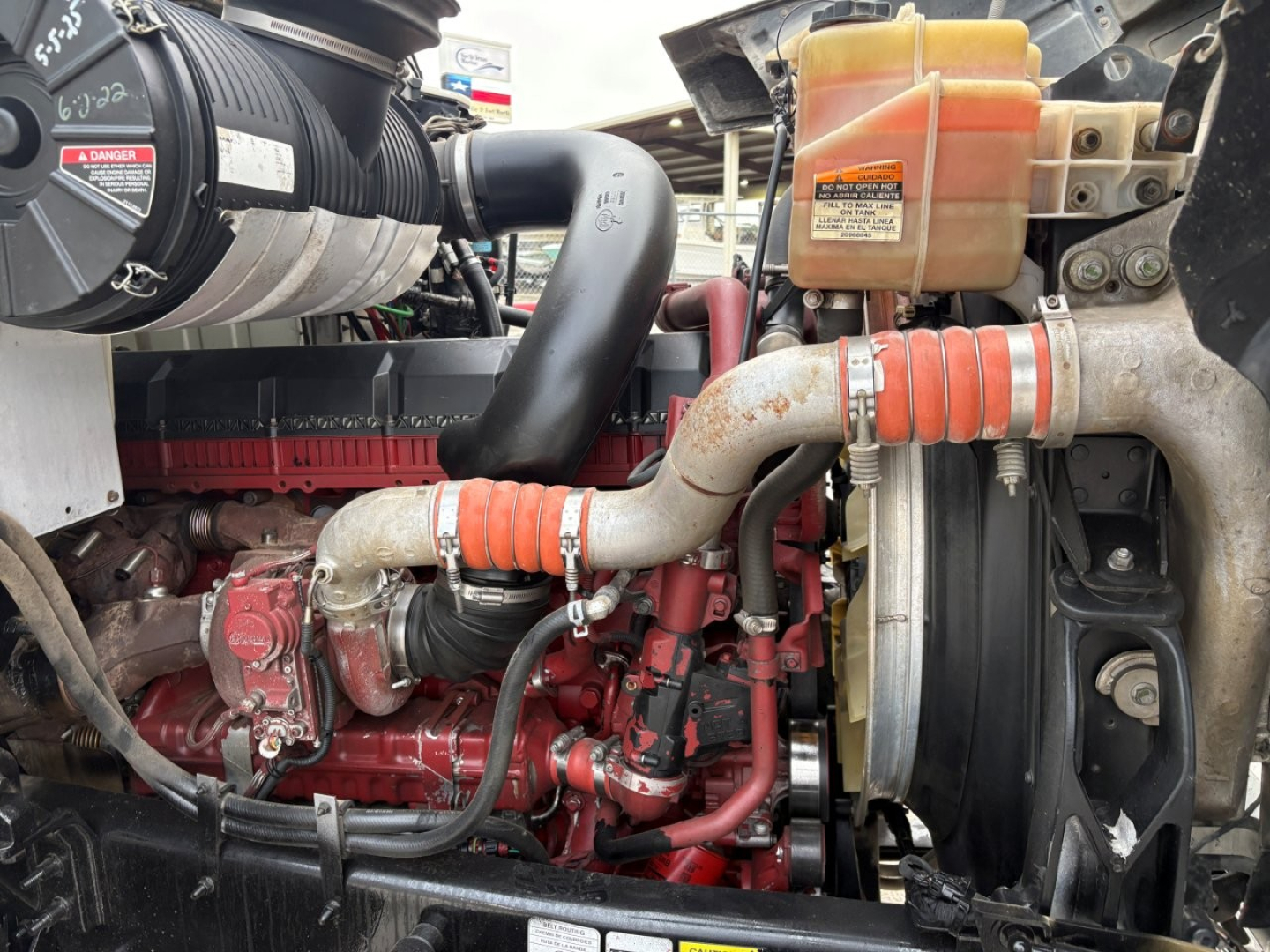 Mack PI64T engine bay showing red engine block, alternator, hoses, radiator, and coolant reservoir with orange cap.
