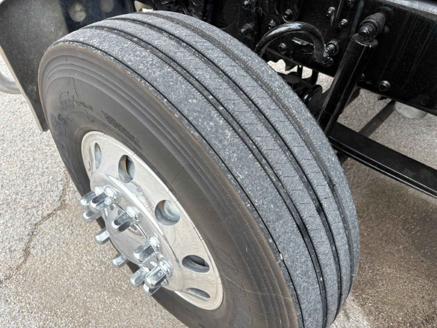 Truck wheel and part of suspension seen from front left, with dual rear wheels and a shiny alloy wheel hub; tread pattern visible on tire; some mechanical components in background.