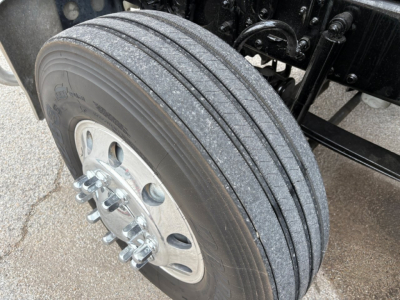 Truck wheel and part of suspension seen from front left, with dual rear wheels and a shiny alloy wheel hub; tread pattern visible on tire; some mechanical components in background.