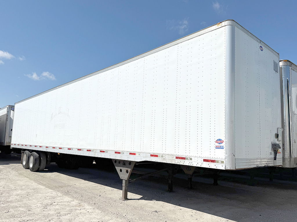 Utility trailer 4000D-X side view showing tandem axles and support legs, white aluminum exterior with rivets and yard backdrop.