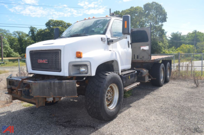 GMC flatbed truck with white cab and dual rear axles, front bumper guard, large tires, and flatbed cargo area visible.