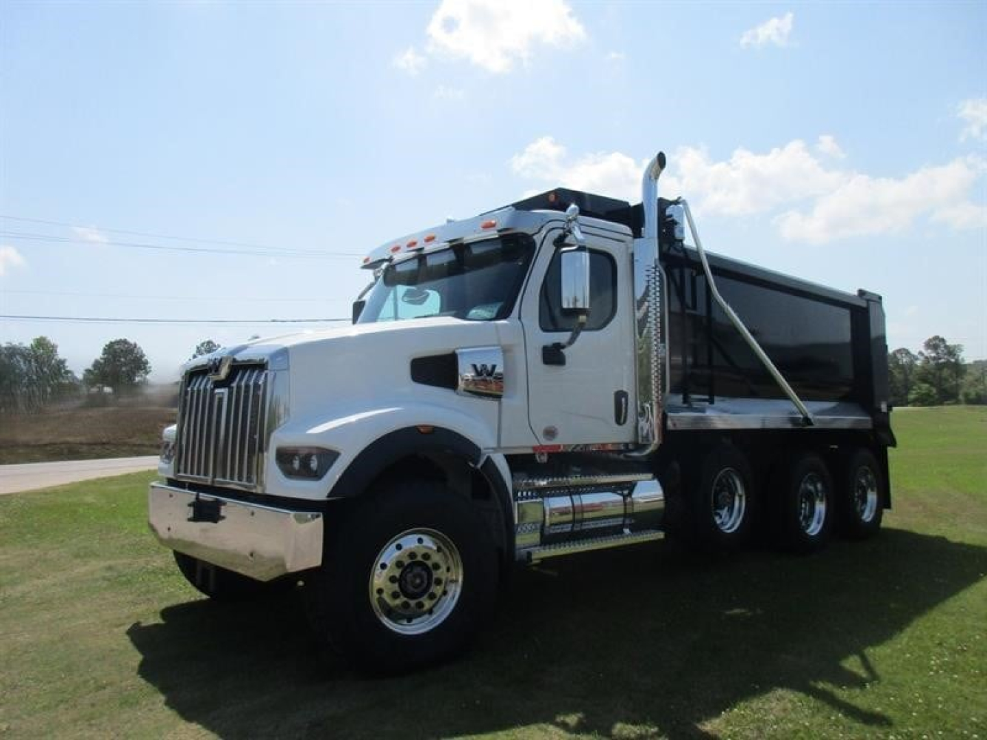 White Western Star dump truck with black dump body, front grille and bumper; side mirrors; three rear axles; chrome exhaust stack; step rails; wheel wells; stock number plate area visible.
