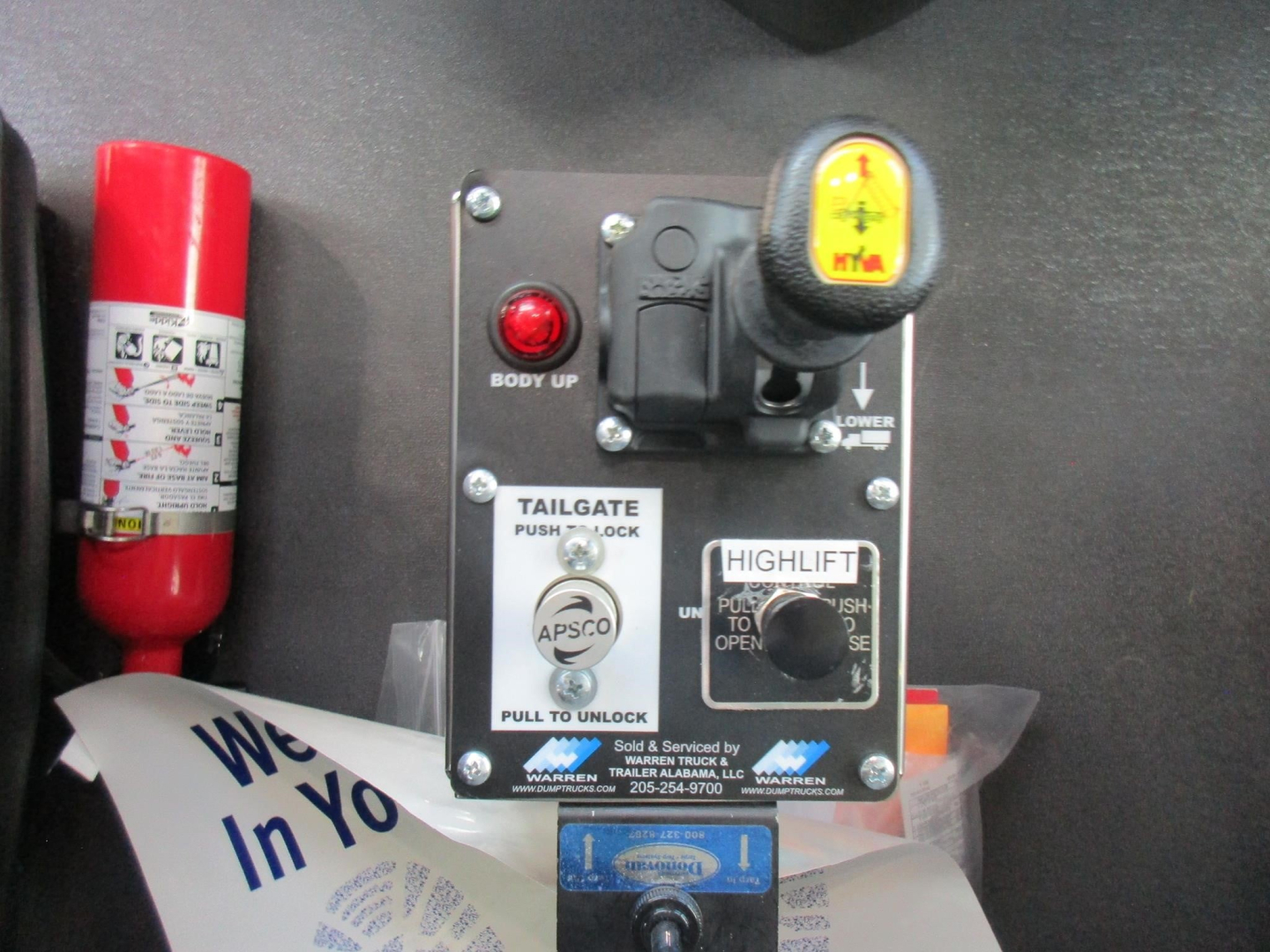Panel showing taillift controls with labels: BODY UP, LOWER DOWN, TAILGATE PUSH TO LOCK, PULL TO UNLOCK, HIGHLIFT, APSCO; a yellow handle with a button on top; a red fire extinguisher on left; warning text plate at bottom.