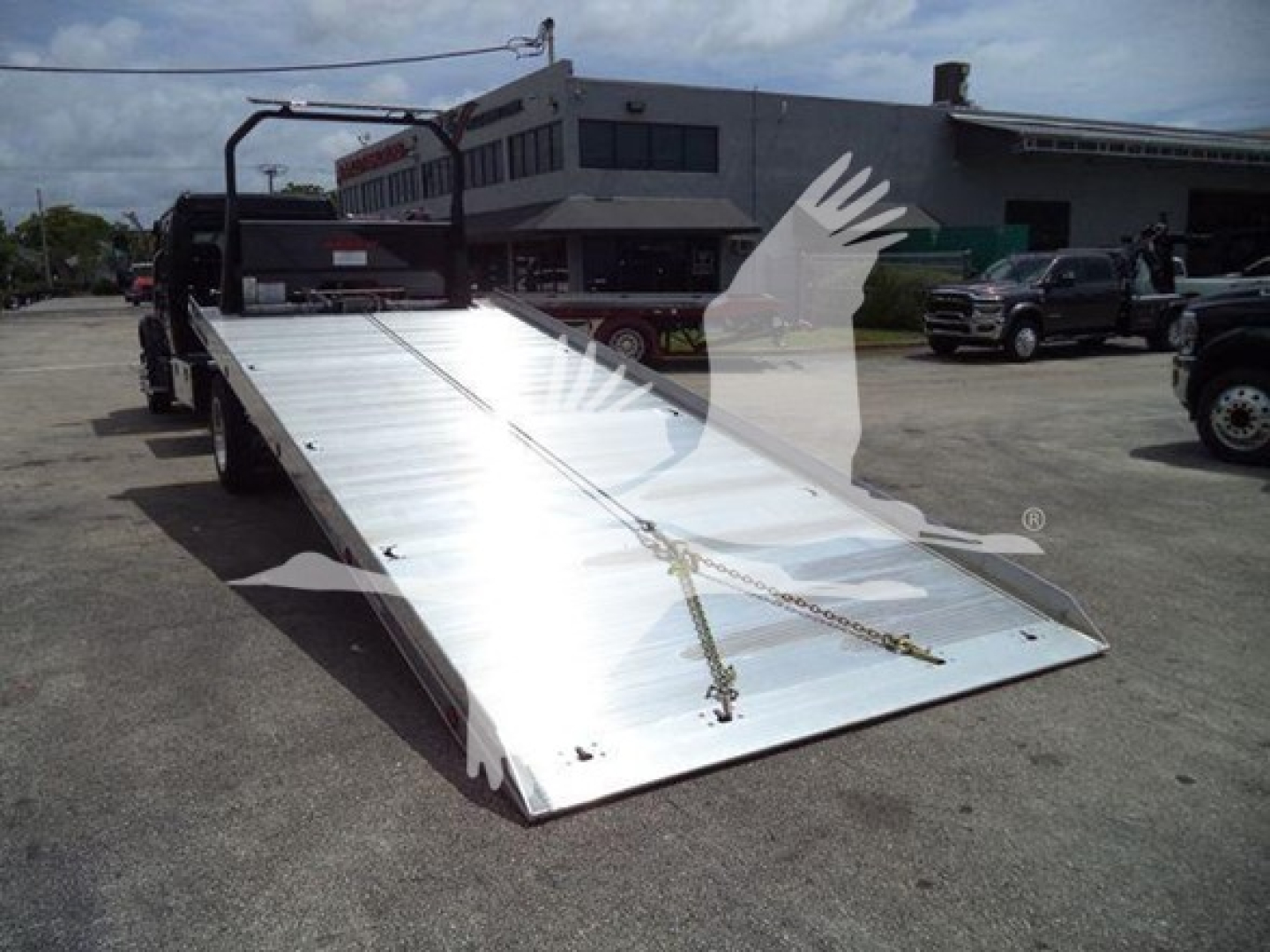 Flatbed tow truck trailer with metal ramp in a dealership lot, Freightliner M2 106 in listing, shows ramp and side rails.