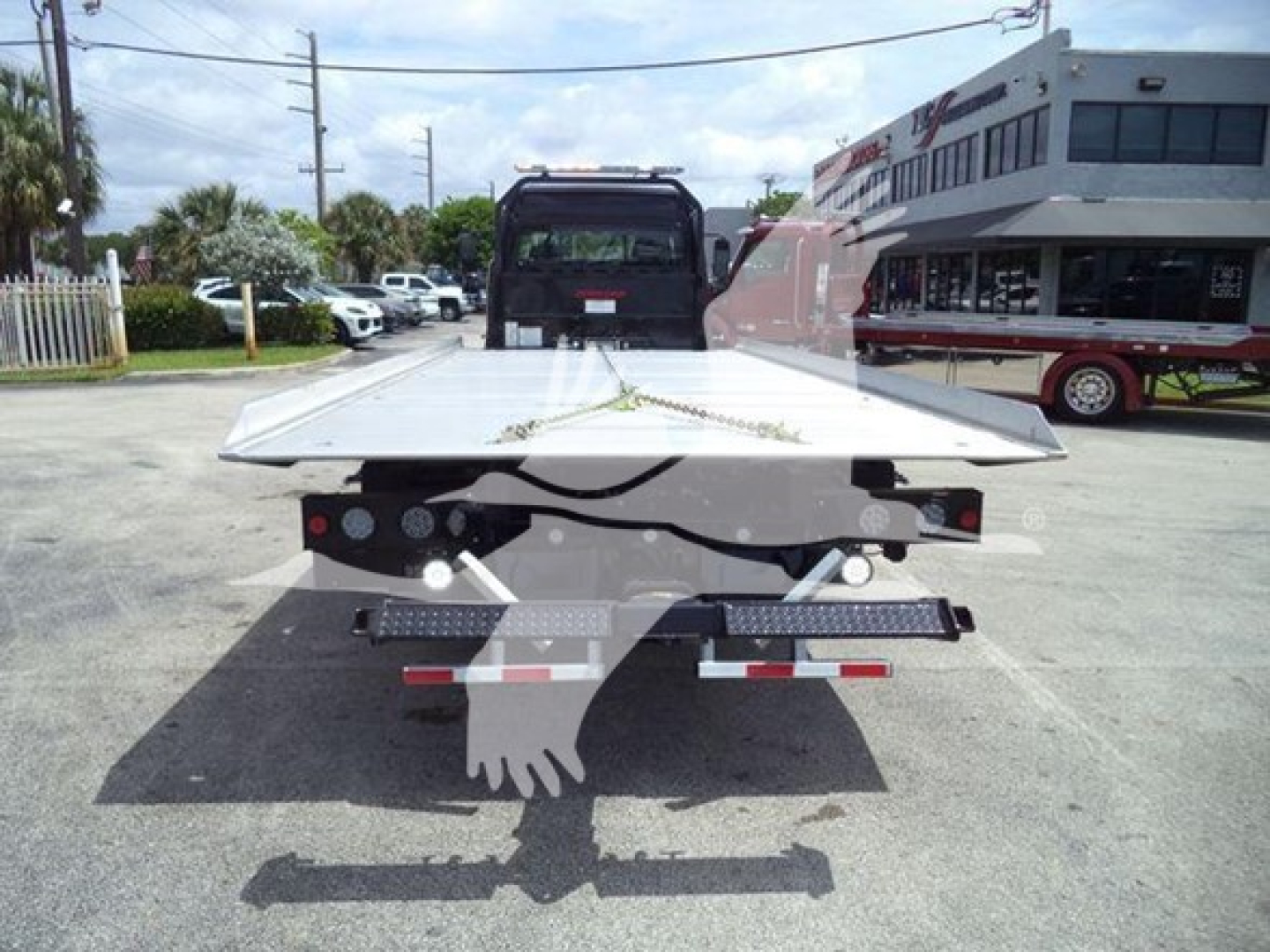 Flatbed tow truck rear view with platform and hydraulic ramps, Freightliner M2 106 dealer lot in background; no legible text on the equipment visible.