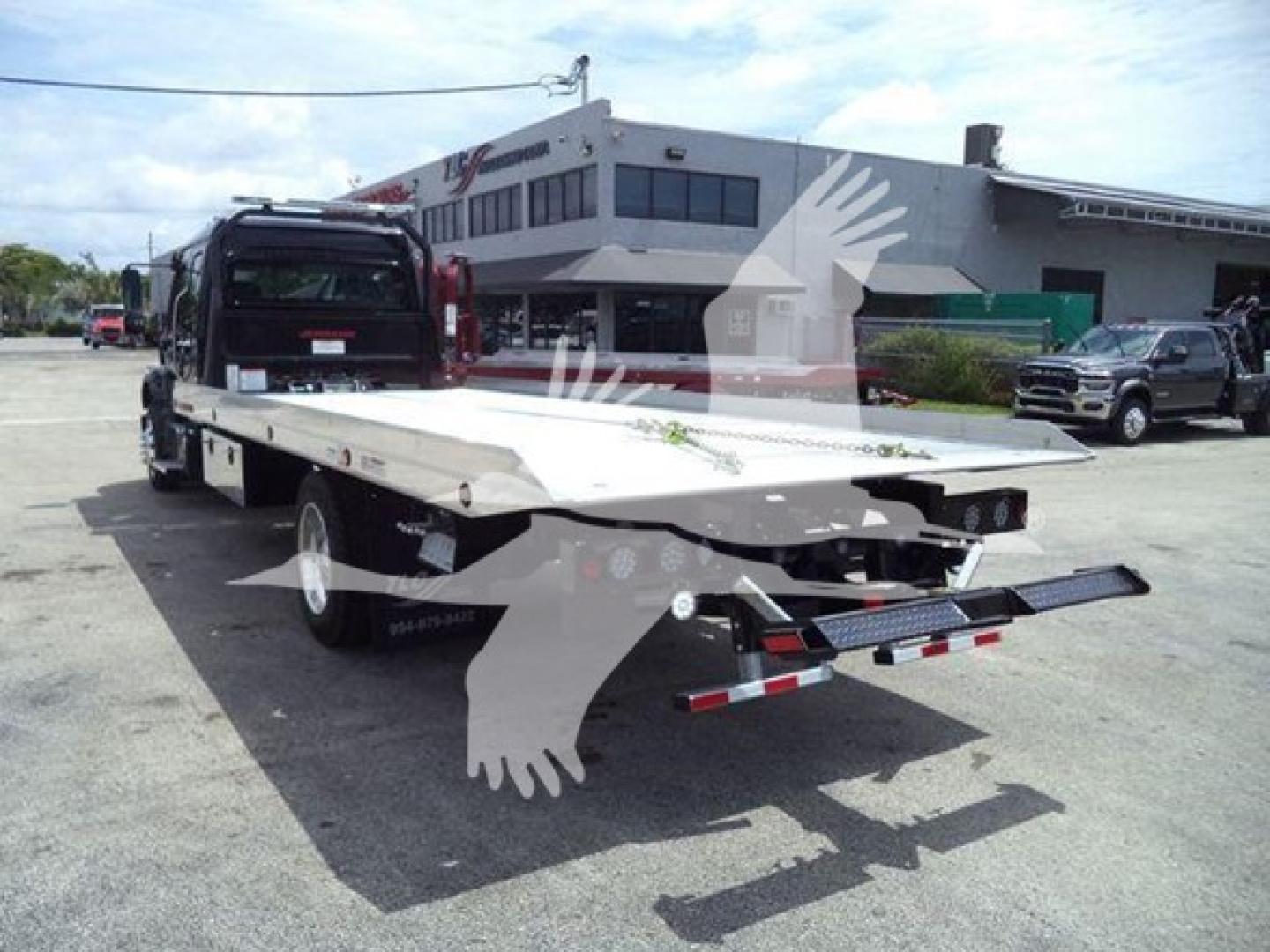 Freightliner M2 106 rollback truck flatbed with rear loading ramps extended, black cab, white bed, dealership lot in background.