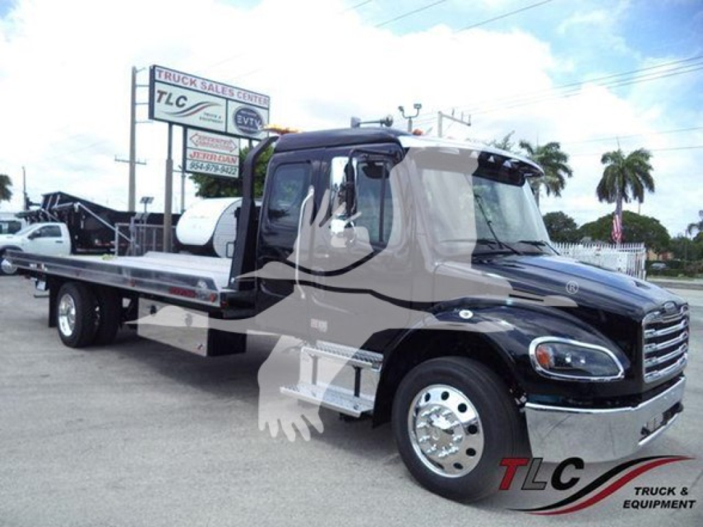 Flatbed truck in a dealership lot with CDL-style cab, black and chrome front, and a long flatbed. TLC Truck & Equipment logo watermark on lower right. Visible text on sign in background includes 'TRUCK SALES CENTER' and 'TLC' signs.