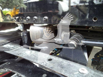 Truck chassis area showing metal frame, hydraulic lines, and mounting brackets in a Freightliner M2 106. Visible bolts and a guard with a shield emblem on a component.