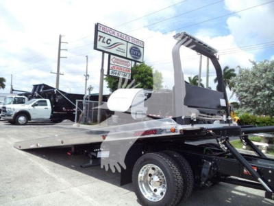 Flatbed rollback tow truck chassis with a raised crane arm, Freightliner M2 106, front wheel visible, dealership sign in background.