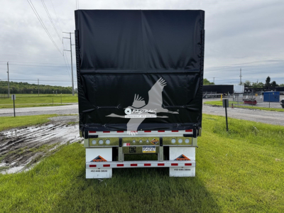 Rear view of a curtain-side trailer with black tarp, RK-100 style, showing rear lights, license plate area, and white curb chain. Brand logo on tarpaulin, license plate visible.