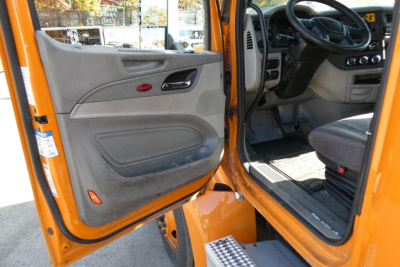 Peterbilt 579 cab interior view with open door showing gray door panel and dashboard, orange exterior, entry steps, and driver seat.