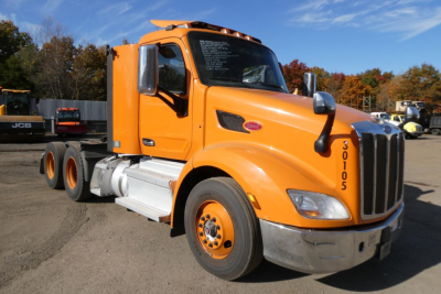 Peterbilt 579 conventional daycab truck in orange, front-left view, showing cab, chrome grille, large side mirrors, and 3-axle rear chassis.