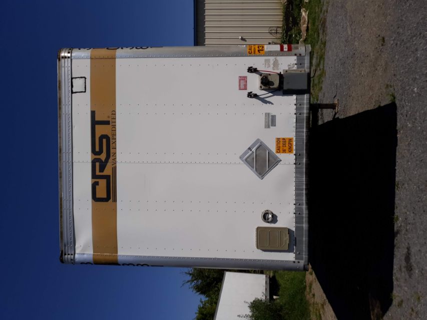 Rear view of a white CRST cargo trailer with a tan stripe and CRST VAN EXPEDITED logo. Visible safety placards, a diamond-shaped reflective emblem, and rear doors.