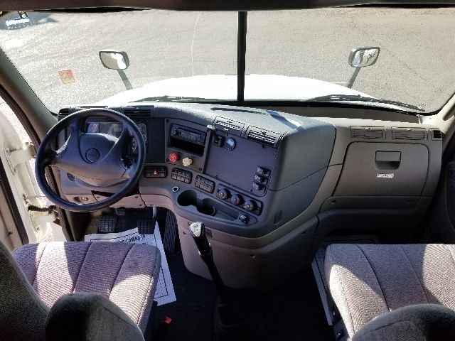Interior of a Freightliner Cascadia truck cab showing steering wheel, dashboard with controls, and gear shifter. Driver seat on left, passenger seat on right, windshield, mirrors visible.