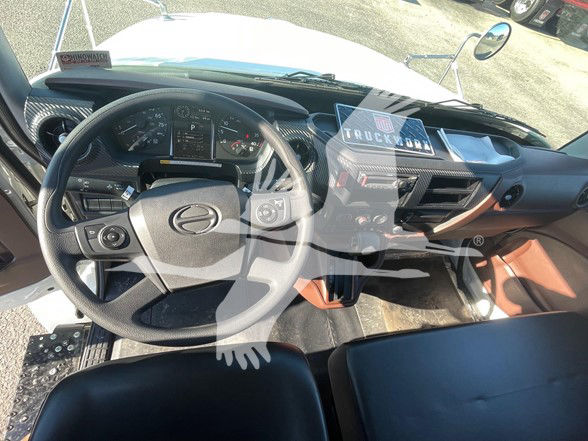 Cab and dashboard of a 2025 Hino L6 truck interior with steering wheel, instrument cluster, center console, and overhead view showing a vehicle info sticker.