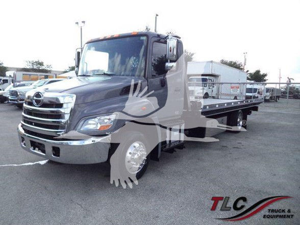 Truck in a lot showing a large gray Hino 258ALP flatbed tow truck with chrome front bumper and grille, side mirrors, and a hydraulic bed. TLC Truck & Equipment logo watermark.