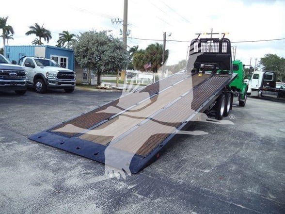 Flatbed tow truck bed on a Freightliner BUSINESS CLASS M2 106 with ramp extended, showing metal decking and wheel lift in a dealership lot.
