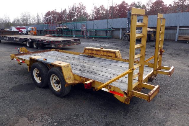 Trailer showing a flatbed with dual axles and a yellow ramp/wing structure at the rear, worn wooden deck, and support frames.