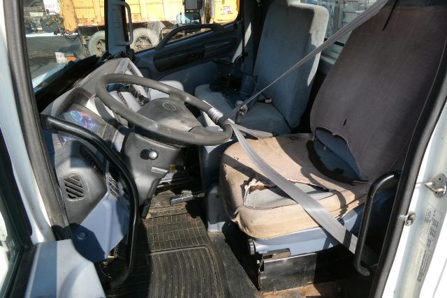 Cab and chassis interior with steering wheel, driver's seat, and passenger seat; seats show wear; visible seat belts; dashboard and floor mat; external door frame visible.