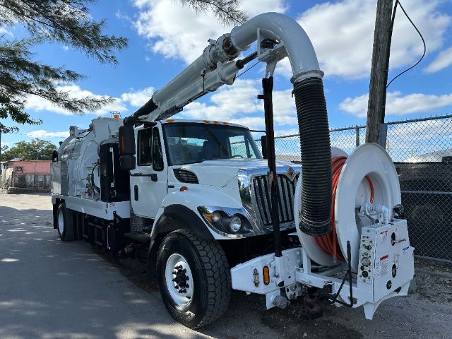 International vacuum truck with a large reel and hose; white cab and pump unit, hose reel on front left, intake hose overhead, blue sky with clouds.