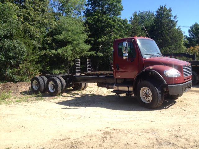 Freightliner M2106 cab and chassis truck with red cab, exposed frame, dual rear axles on dirt lot.