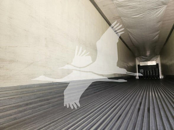 Interior of a refrigerated trailer with ribbed metal floor and empty walls; at far end a metal door with a vertical vent; a watermark of a flying bird overlay.