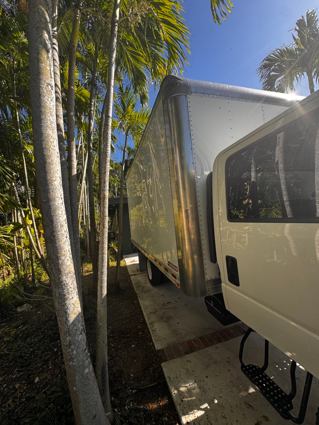 Isuzu NQR box truck side view showing aluminum cargo box with rounded chrome corner post and entry door, white cab, palm trees in background.