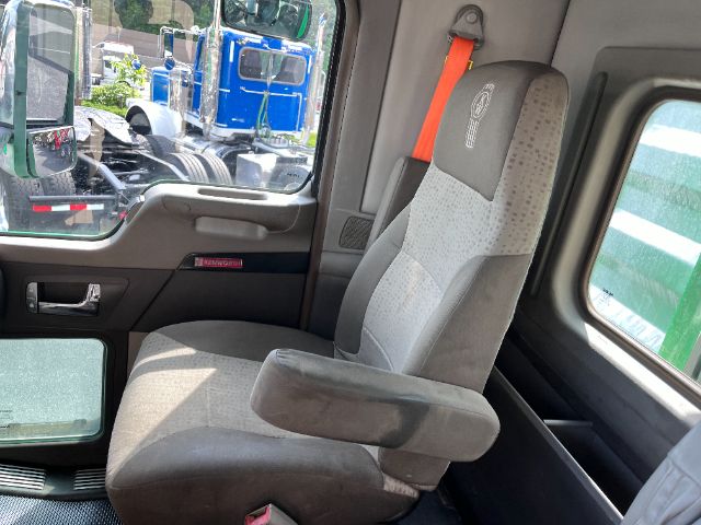 Truck cab interior showing driver's seat with gray upholstery, orange seat belt, and window view of other trucks outside.